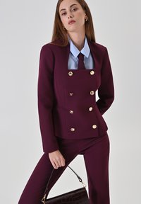 Bordeaux op maat gemaakte blazer met gouden knopen, gecombineerd met een blauwe dress shirt en bijpassende broek. Model houdt een zwarte handtas met een getextureerde afwerking.