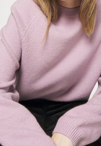 Pull en maille violet clair avec un col rond classique, des poignets côtelés et une texture douce. Présente une coupe décontractée et des détails minimaux.