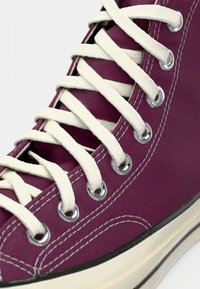 Artimo planu pavaizduotas purpurinis drobės aukšto stiliaus sportinis batelis su baltais raišteliais, sidabriniais akių kabliukais, baltais siūlais ir švelniai balta guma padais su juodu juostele.