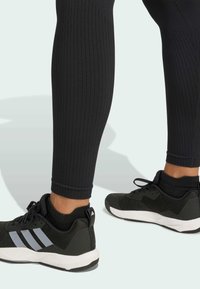Leggings noires côtelées associées à des chaussures de sport noires avec des accents gris et un design texturé. Les chaussures ont une semelle blanche avec un logo.