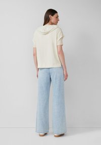 Frau, die seitlich steht, trägt einen kurzärmeligen, cremefarbenen Hoodie und lockere, hellblaue Jeans mit braunen Schuhen vor einem einfarbigen Hintergrund.