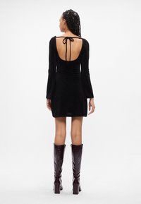 Abito nero a maniche lunghe con schiena scoperta e chiusura a fiocco. Presenta una silhouette aderente e maniche svasate. Abbinato a stivali lucidi marroni al ginocchio.