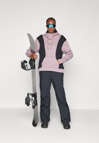Hoodie v svetlo vijolični in črni barvi z blokiranjem barv ter s kabelčki, v kombinaciji s črnimi snežnimi hlačami. Model drži snežno desko z vezmi.