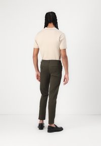 Beige polo paita lyhyillä hihoilla yhdistettynä vihreisiin kapeaan istuvaisiin housuihin ja mustiin loafereihin. Takakuva näyttää räätälöidyn istuvuuden ja ompeluyksityiskohdat.