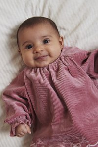 Šypsantis kūdikis guli ant baltos medžiagos, vilkintis ilgomis rankovėmis rožinės kordurojo suknelės su siuvinėta apačia ir raukšlėtu kaklu.