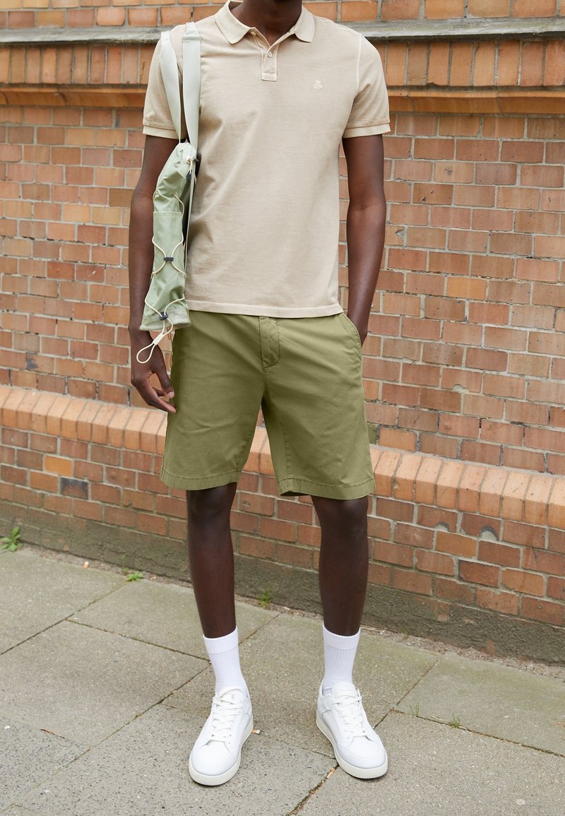 Person som bär en beige polo, olivgröna shorts, vita strumpor och vita sneakers står bredvid en tegelvägg med en vikt grön jacka över ena axeln.