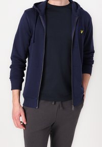 Sudadera con cremallera de color azul marino, con un bolsillo frontal y cordones, combinada con una camiseta de cuello redondo azul oscuro y pantalones de chándal grises.