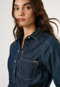 Tume sinine denim-särk, millel on nööpidega sulgemine, klassikaline kaelus, eesmine tasku ja kontrastsed oranžid õmblused.