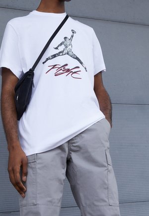 Person iført hvid T-shirt med sort-hvidt billede af en springende basketballspiller og rød tekst "Flight", lysegrå cargobukser og sort crossbody-taske.