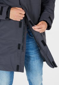 Donkergrijze gewatteerde jas met een rits aan de voorkant en klittenbandsluitingen. Voorzien van ribgebreide manchetten en een gladde stofstructuur. Draagt over blauwe jeans.