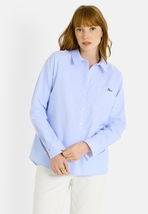 Camisa azul claro abotonada con mangas largas, bolsillo en el pecho con texto bordado y cuello clásico. Detalle a rayas en contraste en el interior de los puños.