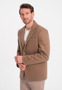 Bruine wolmix blazer met een opstaande revers, enkele knop en een borstzak. Gecombineerd met een lichtgekleurde polo. Glad textuur.