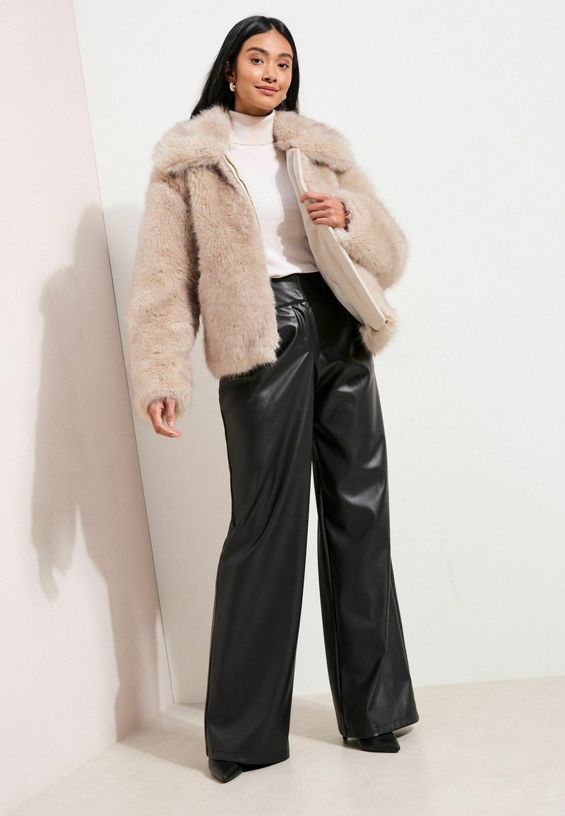 Vrouw in een beige nepbonten jas over een witte coltrui, zwarte wijde leren broek en met een beige clutch in de hand.