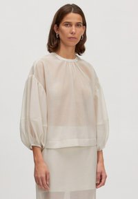Bluse aus transparenter Cremefarbe mit Puffärmeln, Rundhalsausschnitt und Raffinausführung. Weiche Textur, leichtes Material und lockere Passform.