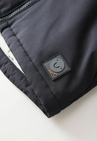 Tissu d'extérieur noir avec une texture lisse, présentant une étiquette rectangulaire avec un visage souriant et le mot " Heureux " en orange.
