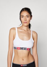 Hvit sports-BH med bred svart elastisk bånd som har rød "DIESEL" logo. Glatt stoff, scoop utringning og tynne skulderstropper.