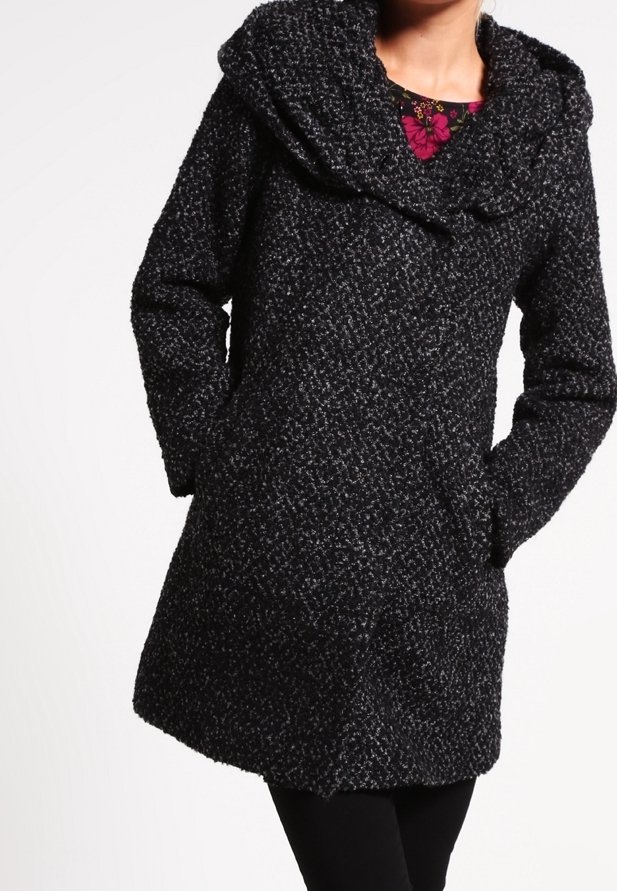 Femme portant un manteau en laine noir texturé avec un grand col, les mains dans les poches, et un haut floral rose visible au niveau du cou.