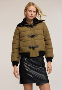 Olivově zelená prošívaná puffer bunda s černým límcem, vybavená uzávěrem na šňůrky a žebrovanými manžetami. V kombinaci s černou koženou sukní.