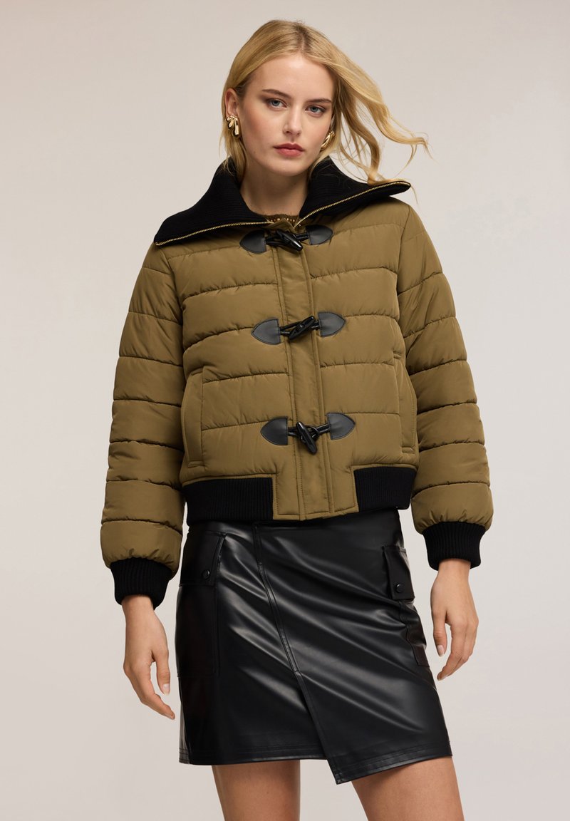 Olivově zelená prošívaná puffer bunda s černým límcem, vybavená uzávěrem na šňůrky a žebrovanými manžetami. V kombinaci s černou koženou sukní.