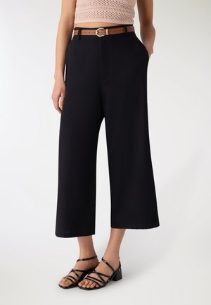 Donna che indossa pantaloni neri a gamba larga e corti con una cintura marrone e sandali neri con tacco largo e cinturino, in piedi davanti a uno sfondo bianco.