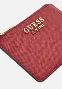 Red textured leather wallet with gold GUESS logo and "EST 1981" embossed, featuring a gold zipper clasp.