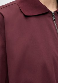 Lähivõte martsipunase lukuga riideesemest, millel on tekstuuriga krae ja hõbedane lukk paremal küljel.