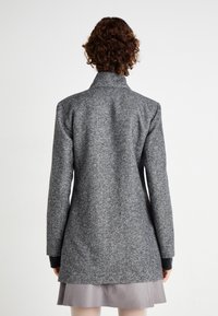 Manteau en mélange de laine gris avec un col montant, des manches longues avec des accents noirs et une coupe longueur genou. La texture semble douce et légèrement structurée.