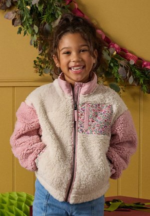 Smilende barn med krøllet hår iført en lyserød og hvid fleecejakke med blomstret lomme, stående indendørs foran en gul væg med grønne dekorationer.
