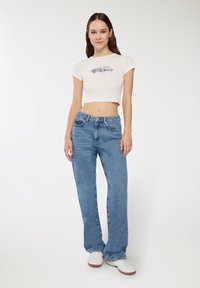 Przycięta biała koszulka graficzna z wzorem samochodu, w połączeniu z jasnoniebieskimi spodniami z wysokim stanem i białymi trampkami, wszystko na neutralnym tle.