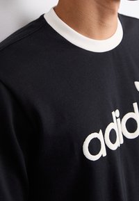 Mustan puuvillan t-paita, jossa on valkoinen ribbineck ja suuri valkoisella brodeerattu logo edessä. Lähikuva kaula-aukosta ja logon yksityiskohdasta.