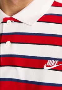 Röd och vit randig polo med marinblå accenter, tillverkad av textil med struktur. Har en vit Nike-logotyp och två knappar vid kragen.