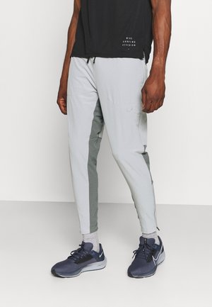 Homme portant un pantalon de sport gris clair avec des panneaux latéraux gris foncé, des chaussures de course noires, des chaussettes gris clair et un t-shirt noir Nike Running Division.