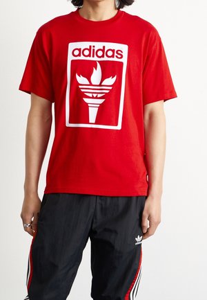 T-shirt en coton rouge avec un grand logo adidas blanc et un motif de flammes dans un contour rectangulaire, accompagné d'un pantalon noir avec des accents rouges.