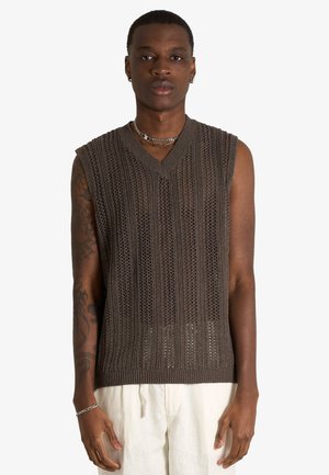 Giovane uomo con capelli corti che indossa un maglione marrone senza maniche e pantaloni bianchi, in piedi con le braccia rilassate ai lati.