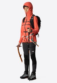 Chaqueta impermeable roja con capucha ajustable, mochila negra, pantalones grises y equipo de senderismo que incluye bastones y herramientas, todo contra un fondo neutro.