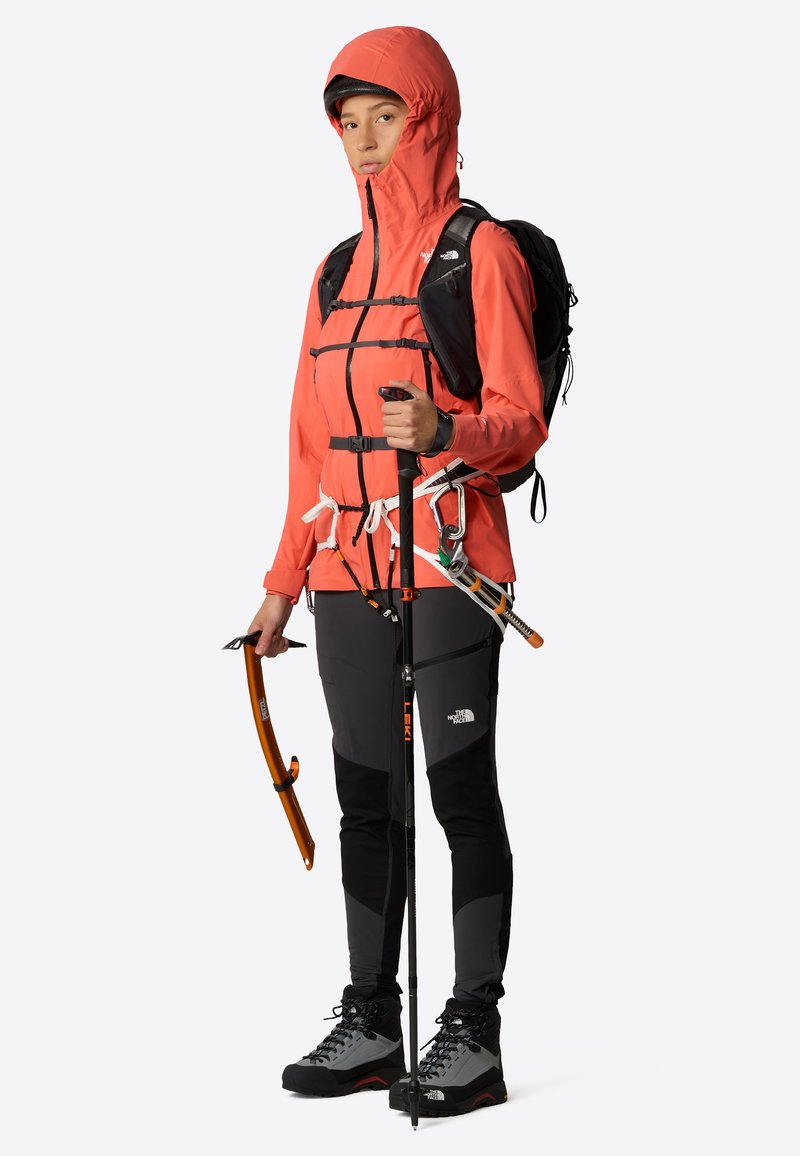 Chaqueta impermeable roja con capucha ajustable, mochila negra, pantalones grises y equipo de senderismo que incluye bastones y herramientas, todo contra un fondo neutro.
