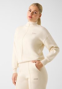 Cremefarbener Zip-up-Sweatshirt aus weichem Stoff mit hohem Kragen und dem "GUESS"-Logo in silberner Stickerei auf der Brust.