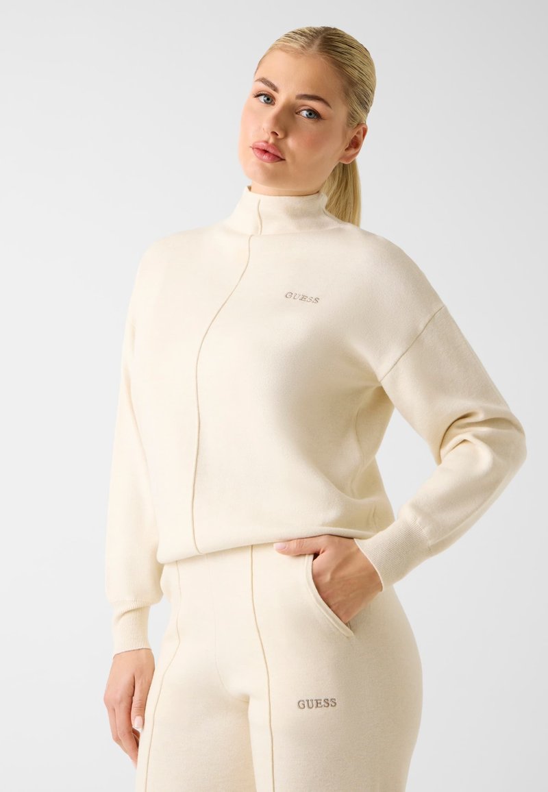 Cremefarbener Zip-up-Sweatshirt aus weichem Stoff mit hohem Kragen und dem "GUESS"-Logo in silberner Stickerei auf der Brust.