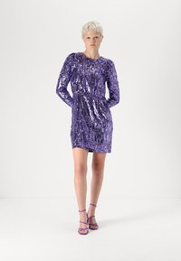 Vestido roxo de lantejoulas com mangas compridas, cintura justa e comprimento acima do joelho. Modelo usa saltos roxos com tiras combinando. Fundo simples.