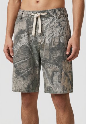 Man wearing gray and beige camouflage-patterned shorts with a white drawstring tied at the waist, standing against a plain background.