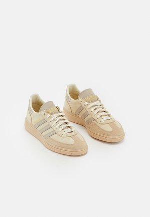 Light beige sneakers with suede and fabric materials, featuring three gray stripes, textured accents, and a tan rubber sole. Laces are white.