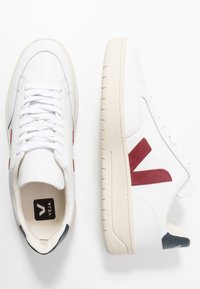 Zapatillas de cuero blanco con un logo rojo en forma de V, cordones planos y una suela de goma beige con un patrón texturizado. Detalle azul marino en el talón.