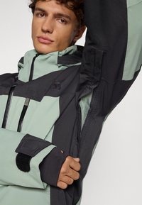 Jachetă izolată cu un design bicolor, mint green și negru. Include buzunare cu fermoar, manșete ajustabile și o căptușeală din plasă.