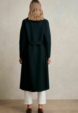 Asmuo su peties ilgio šviesiais plaukais, vilkintis ilgą juodą apsiaustą su diržu, baltas kelnės su apykaklėmis ir rudos kulninės avalynės, stovintis ant smėlinės spalvos grindų.