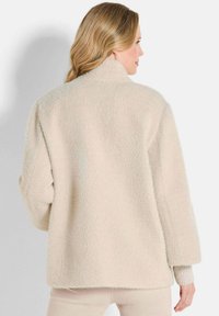 Beige oversized jas van een textiele, zachte stof. Kenmerkt zich door een hoge kraag en ribbelmanchetten. Eenvoudig ontwerp zonder zichtbare hardware.