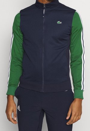Mann trägt eine marineblaue Jacke mit Reißverschluss, grünen Ärmeln und weißen Streifen, kombiniert mit marineblauen Adidas-Hosen.