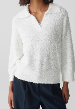 Mujer con suéter blanco texturizado de manga larga y cuello, pantalones azul marino, de pie frente a un fondo gris liso.