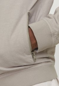 Beige sweatshirt med sidelommer med lynlås, ribbede manchetter og en glat tekstur, der fremhæver et nærbillede af hånden i lommen.