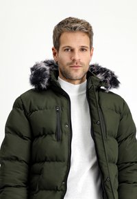 Donkergroene pufferjack met een nepbont capuchon. Heeft ritszakken en een doorgestikt patroon. Draagt over een lichtgrijs trui.