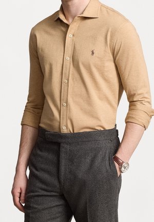 Man wearing a tan buttoned shirt with rolled sleeves, charcoal high-waisted trousers, and a wristwatch with a brown leather strap.